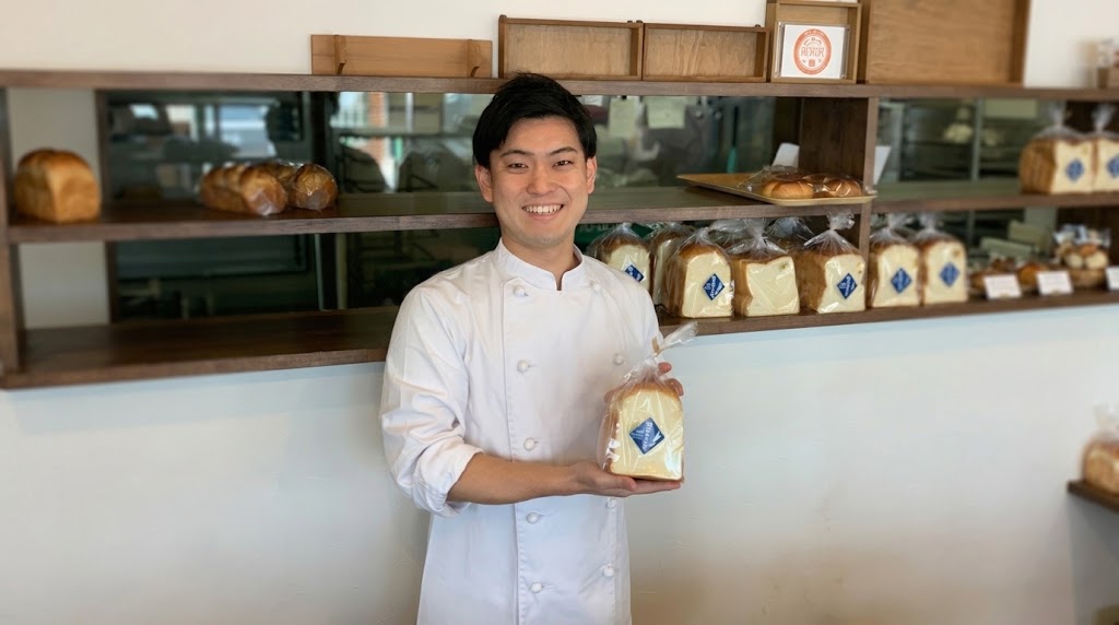 コックコートを着て、食パンを手に店内の棚の前で笑顔を見せる男性
