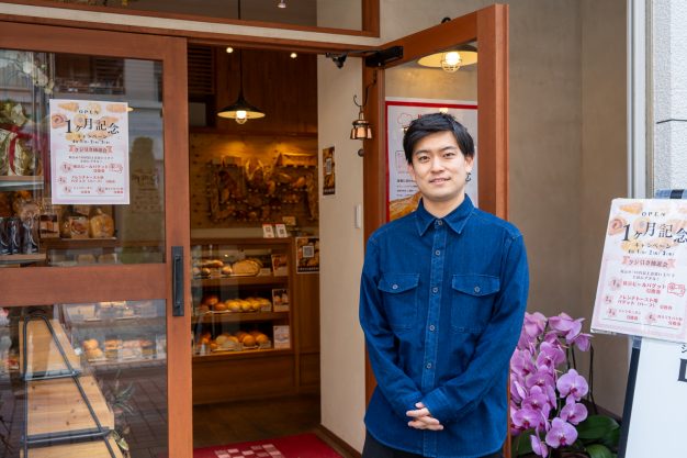 デニムシャツを着て、ベーカリーの店舗入口の前に立つ男性