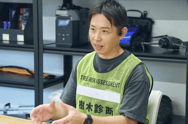 「樹木診断」と書かれたベストを着て、身振りを交えながらインタビューに答える男性